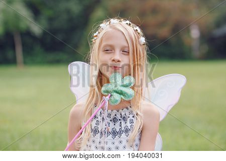 Portrait of smiling blonde white Caucasian child kid girl with long hair wearing pink fairy wings and tutu tulle skirt holding magic wand happy lifestyle childhood game play concept