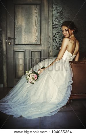 Beautiful bride sits in a leather chair in a dark room in the hands of a wedding bouquet