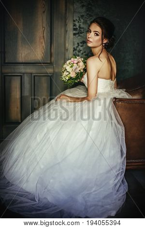 Beautiful bride sits in a leather chair in a dark room in the hands of a wedding bouquet
