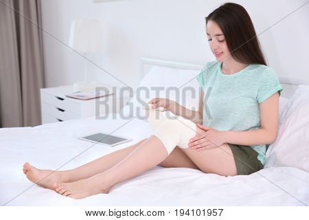 Beautiful young woman applying bandage onto leg while sitting on bed at home