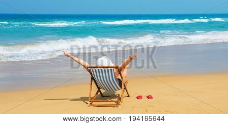 Happy woman in white sunhat on the morning beach sitting on deckchair with hands up.Vacation and travel concept.