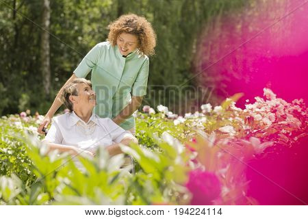 Senior woman on a wheelchair and looking at her young nurse surrounded by flowers