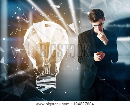 Handsome young businessman using smartphone in blurry interior with glowing geometric light bulb. Digital networking concept. Double exposure