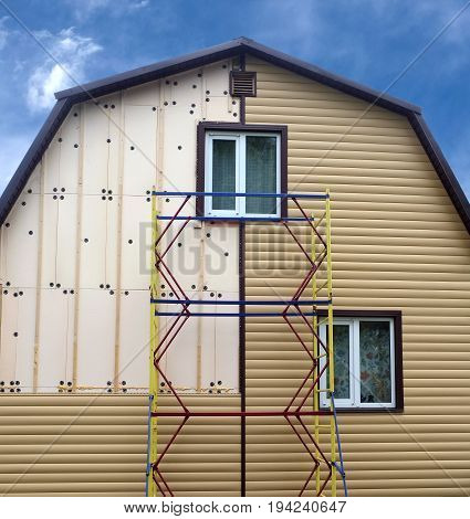 Beige siding panels mounting with scaffolding on beautiful rural house in sunny day front view