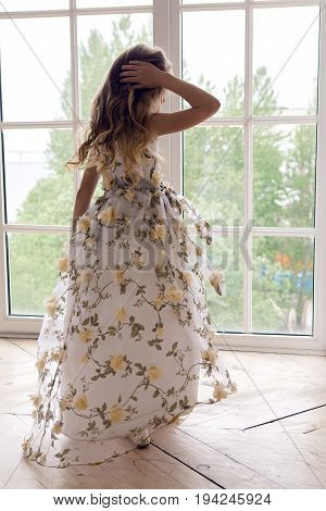 baby girl in a long floral dress stands at a large window