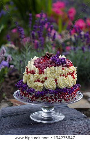 birthday cake with chocolate ganache decorated with roses lavender and berries, garden with flowers in the background