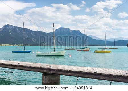 Small sailboats anchoring in alpine lake. Landing stage and mountains framing the boats. Bavaria, Germany, Allgau, Forggensee.