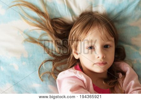 Portrait of a little girl with long hair lying on a sheet with cloudy sky. She is sad and looking at the camera.