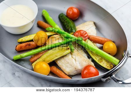 Grilled fish and vegetables with sauce served in metalic bowl on marble serface