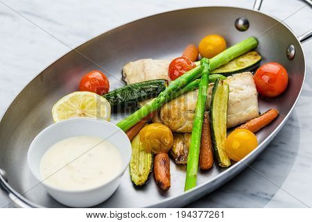 Grilled fish and vegetables with sauce served in metalic bowl on marble serface