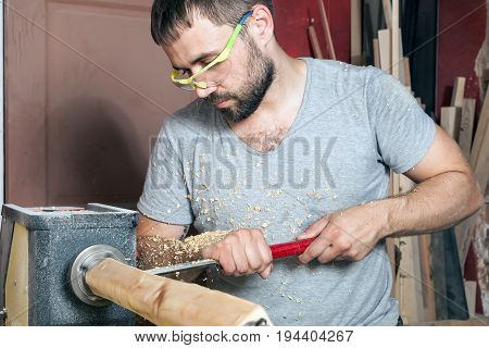 A young brunette man a builder in green protective goggles a gray T-shirt and a beard makes a wooden product on a lathe in the workshop