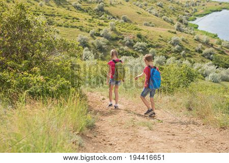 Teen boy and girl with backpacks on the back go hiking, travel, beautiful landscape, hills and pond