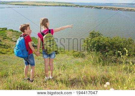 Teen boy and girl with backpacks on the back go hiking, travel, beautiful landscape, hills and pond