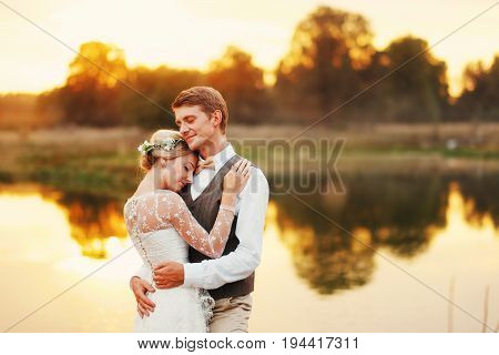 Close-up portrait of a wedding couple against a background of water in the setting sun. Lace dress of the bride