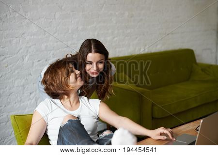 Two brunette girl friends look new modern collection in online shop using internet high wireless connection. Smiling woman with good mood choose dess for romantic meet with boy. Meeting site concept