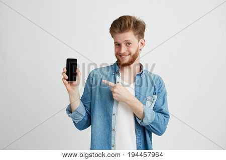 Cheerful young handsome man smiling looking at camera pointing finger at smartphone in his hand over wite background. Copy space.