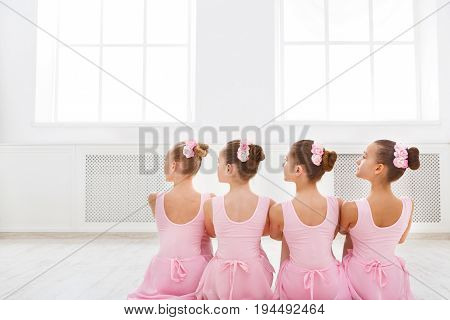 Little ballerinas in ballet studio. Group of girls having break in practice, sitting on floor, back view. Classical dance school