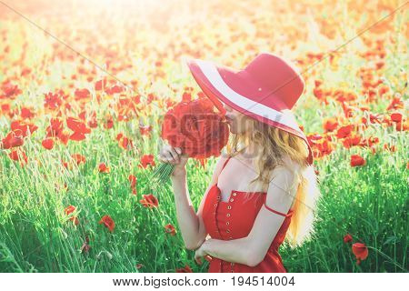 woman with long curly hair in red dress and retro hat hold flower bouquet in field of poppy seed on green stem on natural background summer drug and love intoxication opium pin up woman