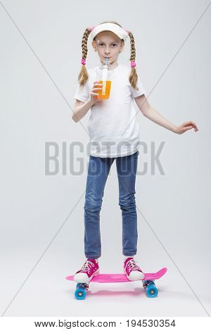 Full Length Portrait of Happy Caucasian Blond Girl in Visor Ballancing on Pennyboard With Cup of Juice.Against White. Vertical Image