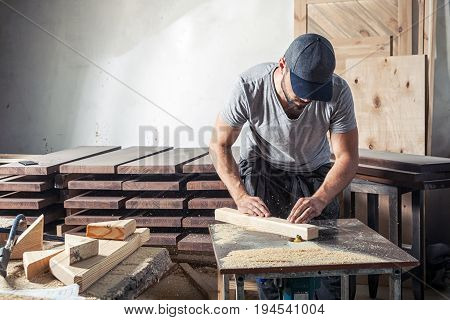 A young man with a cap and a gray T-shirt by profession carpenter builder equals a wooden bar with a milling machine on a wooden table in the workshop around wooden boards