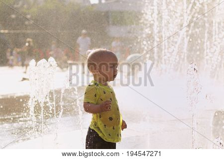 Cute Toddler In Wet Clothes Standing In The Center Of City Fountain. Droplets Of Water Sprayed In Th