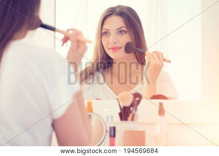 Beauty woman applying makeup. Beautiful girl looking in the mirror and applying cosmetic with a big brush. Girl gets blush on the cheekbones. Powder, rouge
