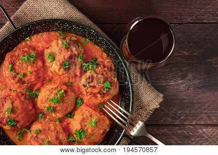An overhead photo of meatballs in tomato sauce, shot from above in the skillet on dark rustic textures, with a place for text, a fork, and a glass of red wine