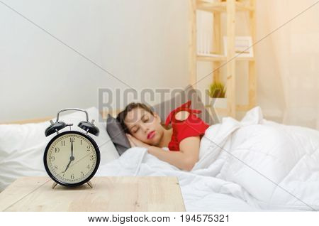 A asian young woman in red pajamas sleeping on bed and alarm clock times at 7 o'clock morning on wooden desk in bedroom at the morning soft focus selective focussunlight effect