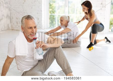 Fitness instructor. Positive delighted elderly woman sitting on the floor on the background and stretching her back while following her instructor