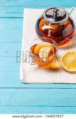 Glass cup of tea with lemon and glass teapot on blue wooden table.
