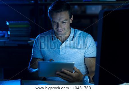 Long hours. Bright diligent hardworking man sitting in the office and reading information on his gadget while completing the task for today