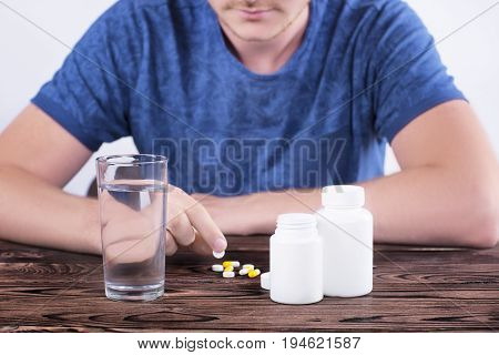 A young guy in the blue T-shirt sits at a wooden brown table and holds a pill in his hand. There is a glass of water on the table, two boxes of medicines and white and yellow tablets on the table.