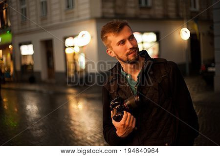 Young male photographer stands on city street in evening. In background there are night city lights. Self-portrait