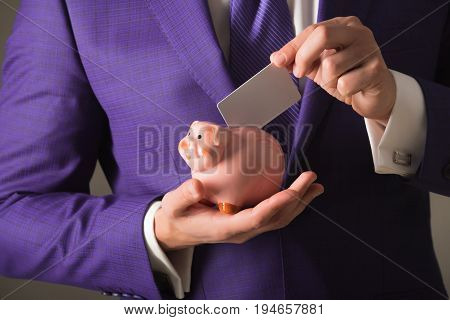 hands putting blank business card in piggy bank for saving on grey background. Stylish formal blue suit and tie. Banking money loan moneybox ecash and information