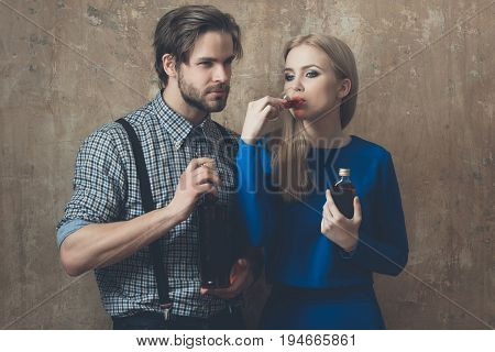 Girl drinking glass of liqueur with man. Pretty woman with blond hair and handsome macho holding bottles with alcoholic drinks on beige wall. Alcohol and convive. Unhealthy lifestyle. Bad habits