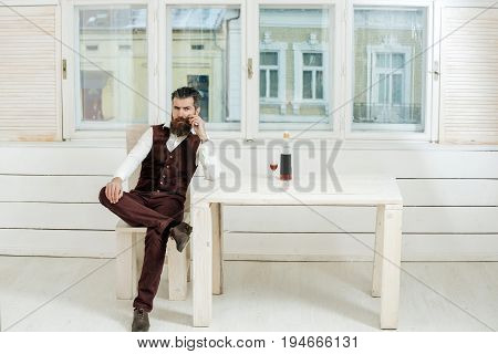 Man with beard and moustache sitting on wooden chair at table with bottle and glass of red wine. Brutal hipster indoors on white window background. Alcohol and convive. Unhealthy lifestyle. Bad habits