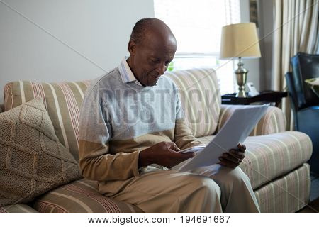 Senior man using phone while holding document in living room at home