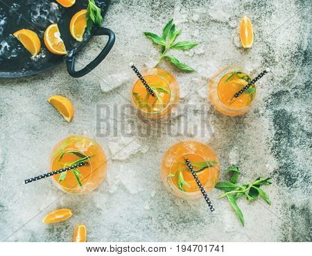 Refreshing cold alcoholic summer citrus cocktail with orange, peppermint and crushed ice in stemless glasses over grey concrete background, top view