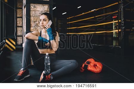 Worn out and serious cute woman with wrapped fists for fighting resting on the floor in gym after effective training and calling her friends on mobile