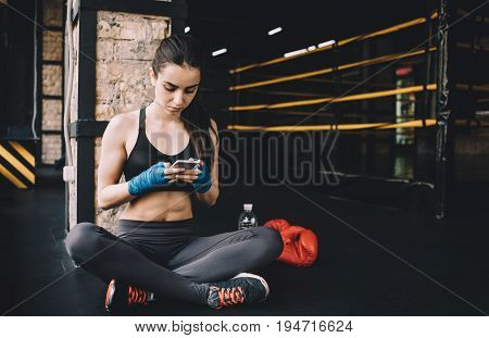 Attractive woman in sportswear sitting near the boxing ring and having some rest. She is scrolling in her mobile phone or chatting with friends
