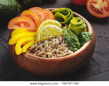 Close up view of vegetarian buddha bowl with green buckwheat, spinach, avocado, tomatoes and yellow sweet pepper paprika on dark concrete background with copy space
