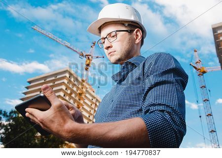 builder is a man in a protective helmet, glasses and a tablet computer in the hands. a crane, a multi-storey building outside, on the street. Concept construction, business, technology