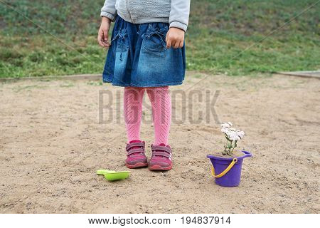 Little girl playing in a sand-box on a cloudy summer day She is planting a flower in a toy bucket. Her face is not seen.