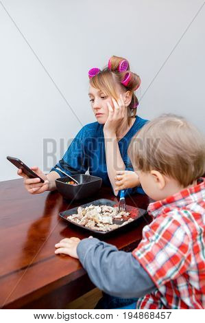 Bored tired white Caucasian young woman mother housewife with hair-curlers in hair looking on phone surfing Internet. Child son boy sitting eating meal lunch. Lifestyle concept.