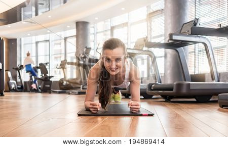 Front view of a young fit woman smiling while practicing the forearm plank exercise for core strength indoors at the gym