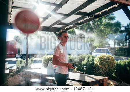 Attractive millennial man with fashionable haircut in trendy outfit stands in line for brunch outside restaurant on terrace with sprinkled water for cooling on hot day
