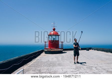Handsome young man in traveler outfit stands near bright red lighthouse sets up timer on smartphone on selfie stick ready to make photo for social media
