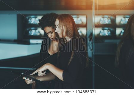 Two young female co-workers looking at something on screen of digital tablet in luxury office young afro american probationer girl consult an beautiful white businesswoman about future presentation