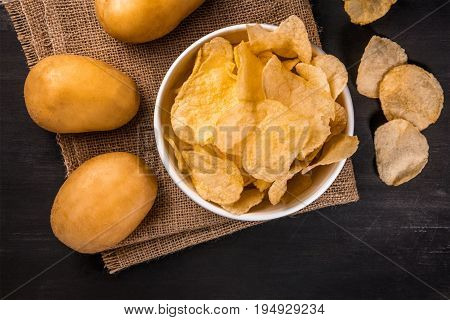 A photo of a bowl of potato chips with potato spuds, shot from above on a burlap and a black texture with a place for text