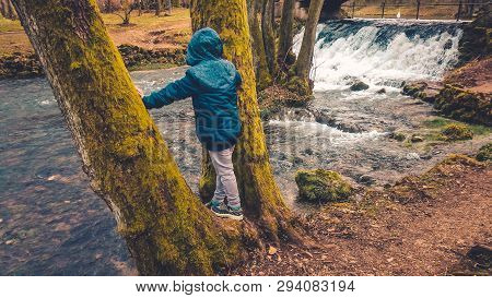 Young Boy Standing On The Tree And Enjoying The Picturesque View Of The River.